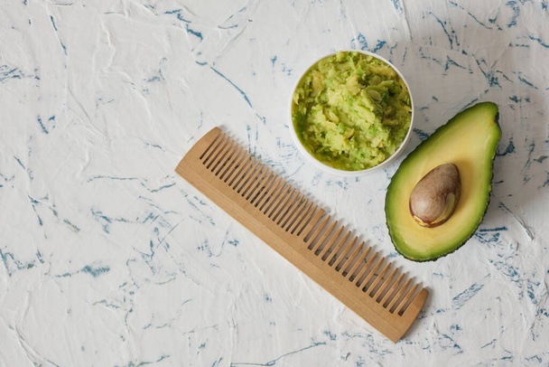white mockup plastic jar with avocado puree, cut in half avocado and wooden comb on light background - Фото, изображение