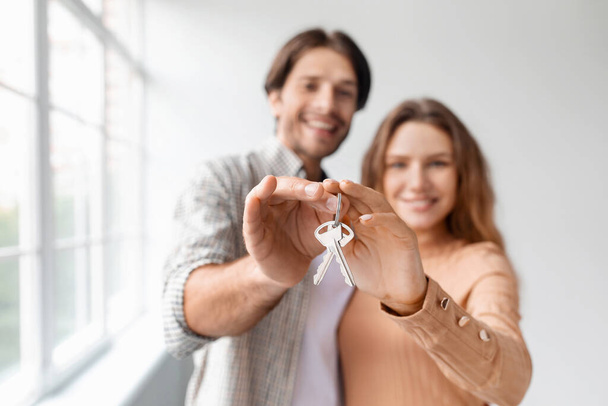 Ευτυχής χαρούμενος καυκάσιος νεαρός άνδρας και γυναίκα σε άδειο δωμάτιο με πανοραμικό παράθυρο, εμφάνιση κλειδιών - Φωτογραφία, εικόνα
