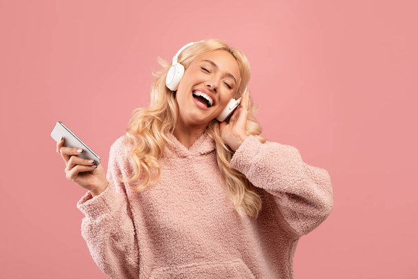 Señora feliz llena de alegría con auriculares y smartphone escuchando música y bailando sobre fondo rosa - Foto, Imagen