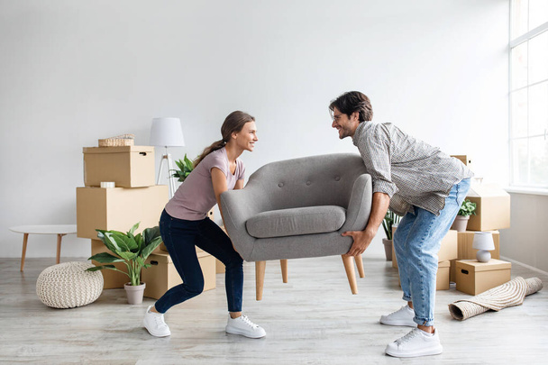 Smiling caucasian millennial wife and husband carry armchair in room with cardboard boxes - Photo, Image