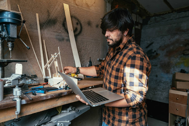 Inżynier używający laptopa do zbadania ramienia robota. Poważny człowiek robi drewniane ramię robota w swoim warsztacie. - Zdjęcie, obraz