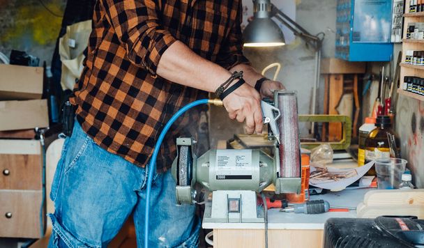 Egy névtelen ács, aki Bench Grinderrel dolgozik a műhelyében. Felismerhetetlen ember tisztítja az elektromos csiszolóját légkompresszorral a garázsában.. - Fotó, kép