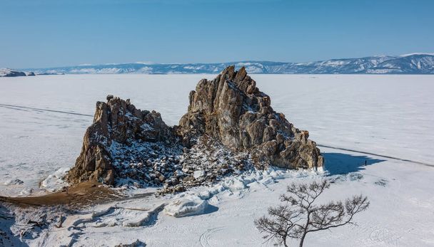 На замерзлому озері здіймається мальовнича двоголова скеля. Сніг на схилах і на льоду. Гірський хребет проти блакитного неба. Байкал. Гора Шаманка - Фото, зображення