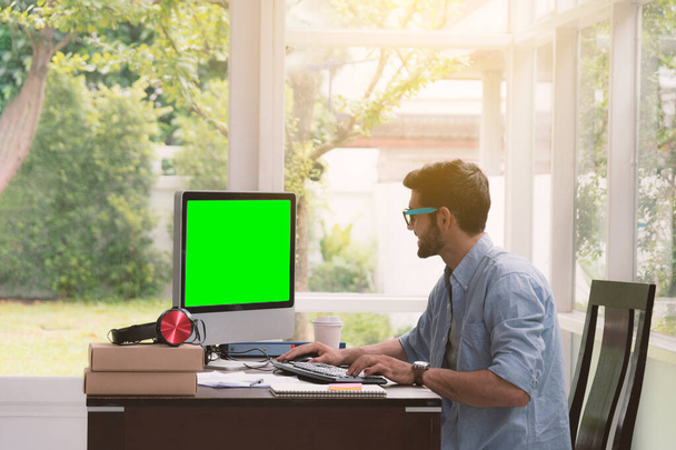 Mann arbeitet abends, wenn die Sonne durch den Raum scheint, am grünen Bildschirm seines Computers im Home Office. - Foto, Bild