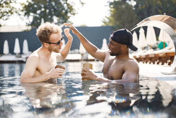 Two male friends relaxing with a cocktails in a swimming pool - Photo, image