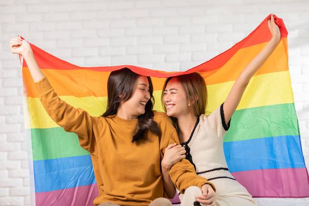 LGBTQ Asian lesbian young couple smiling holding pride flag happiness with love together.Cheerful Homosexual couple embracing and looking at camera under LGBTQ pride flag.LGBTQ Lifestyle Pride Month - Photo, image