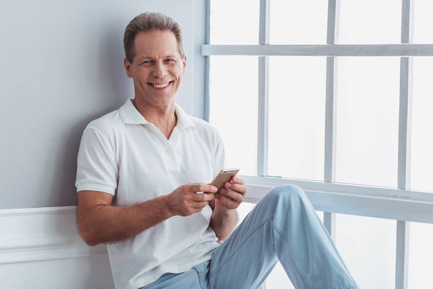 Bello uomo di mezza età in abiti casual sta usando uno smartphone e sorridente mentre seduto vicino alla finestra a casa
 - Foto, immagini