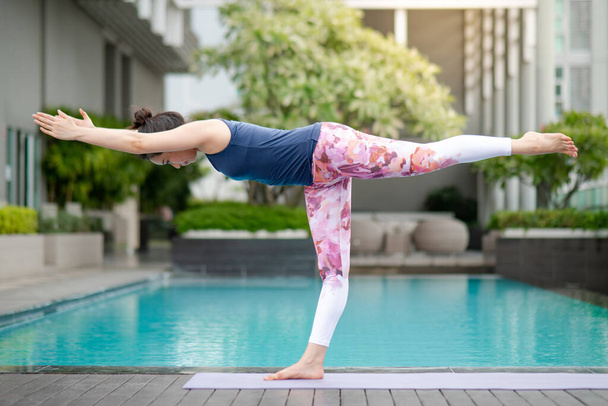 Attractive young Asian woman doing yoga exercise with warrior-III pose or Virabhadrasana III posing near swimming pool. Workout at home. Healthy lifestyle concept. International Yoga Day - Photo, Image