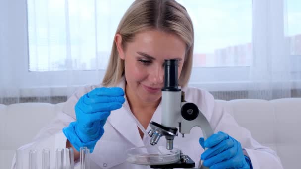 naispuolinen kemisti lääketieteellisessä puvussa ja suojakäsineissä suorittaa tutkimuksia näytteillä, joissa on mikropipetti ja koeputket mikroskoopilla työskentelyä varten laboratoriossa. - Materiaali, video
