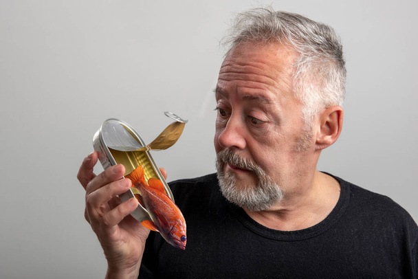 Mężczyzna w średnim wieku otwiera puszkę z rybą w puszce, z której wychodzi żywa ryba. Szare tło - Zdjęcie, obraz
