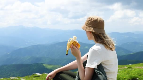 viajero explorador chica come plátano fruta se relaja en la cima de la montaña en un día de verano soleado. Éxito, lujuria errante y concepto deportivo. - Metraje, vídeo