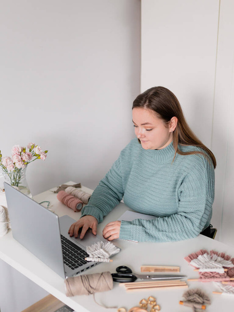 Молодая стильная женщина использует ноутбук дома, смотрит на экран, читает или пишет электронную почту. Женское хобби. Работа над онлайн концепцией исследовательского проекта. Магазин макраме ручной работы - Фото, изображение
