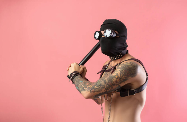bivakmuts man met lederen banden op zijn lichaam voor bdsm seksspeeltjes met een rubberen stokje in zijn handen - Foto, afbeelding