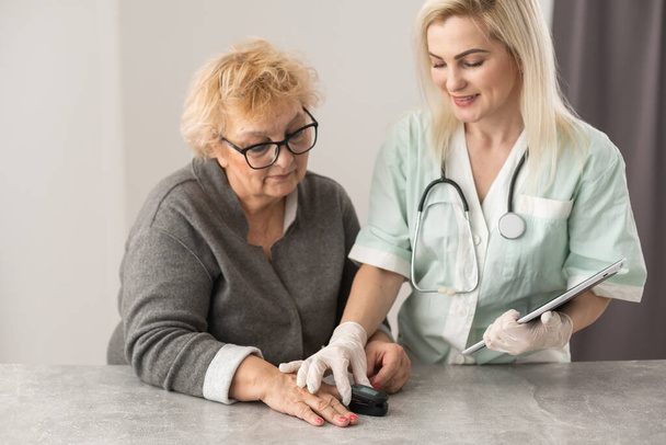 Vrouwelijke verzorger met vingertoppolsmeter, bevestigd aan vinger van ouderen, gezondheidscontrole, meting zuurstofverzadigingsniveau en hartslag thuis tijdens COVID-19 of Coronavirus pandemie. - Foto, afbeelding