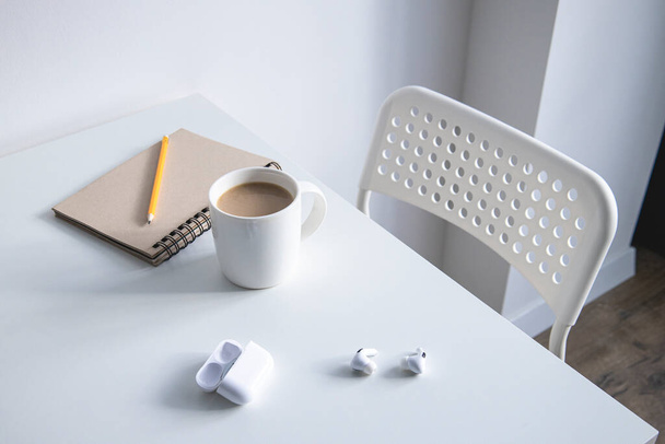 Witte beker met koffie, notitieblok, potlood en draadloze hoofdtelefoon op een witte tafel - Foto, afbeelding
