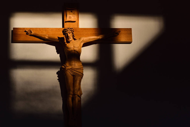 Na ścianie wisi tradycyjny i religijny drewniany krzyż z rzeźbionym Zbawicielem. Światło i cień świecą na krzyżu i ścianie za nim. - Zdjęcie, obraz