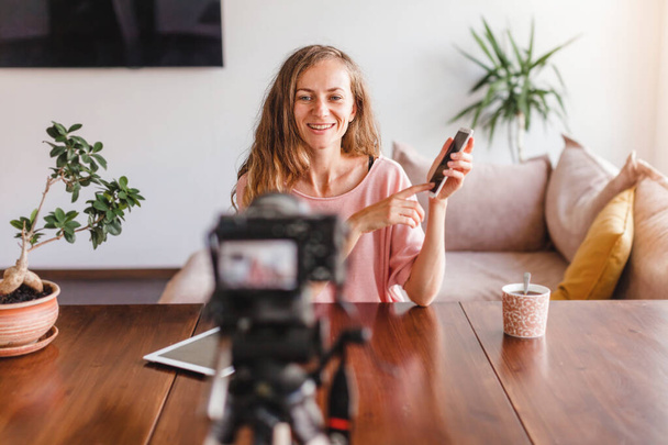 Young attractive happy woman blogger or vlogger looking at camera and talking on video shooting with technology. Social media influencer people or content maker concept in relax casual style at home - Photo, Image