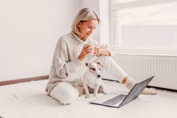 Relaxed middle-aged woman is working on a laptop at her home. Adult female is sitting on the floor near the radiator in a white knitted sweater with her dog and watching movi - Zdjęcie, obraz