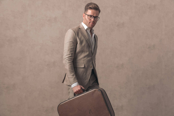 cool young businessman with suitcase walking and being confident on beige background in studio - Fotoğraf, Görsel
