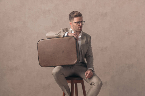homme de mode décontracté intelligent avec des lunettes regardant vers le côté et tenant sac sur la cuisse et assis sur fond beige en studio - Photo, image