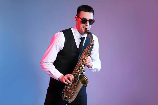 Молодой человек в элегантном наряде играет на саксофоне на цветном фоне - Фото, изображение