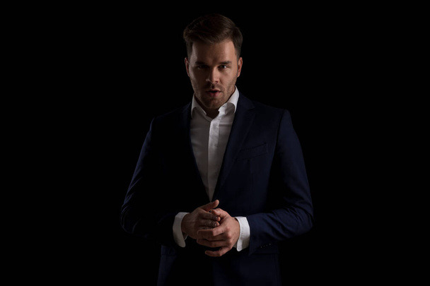 portrait of attractive fashion man in navy blue suit rubbing palms and posing on black background in studio - Fotó, kép