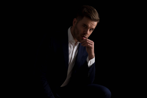 Портрет сексуального бизнесмена в моде легкий трогательный подбородок, думая и будучи соблазнительным, сидя на черном фоне в студии - Фото, изображение