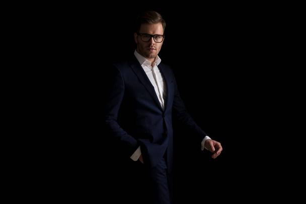 modelo de moda atractiva con gafas de la mano en el bolsillo y caminar sobre fondo negro en el estudio en una luz de moda - Foto, imagen