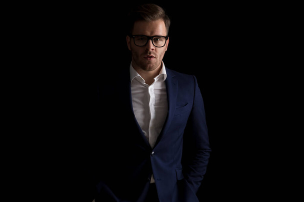 retrato de chico guapo de la moda con gafas cogidas de la mano en los bolsillos y posando en una luz de moda sobre fondo negro en el estudio - Foto, imagen