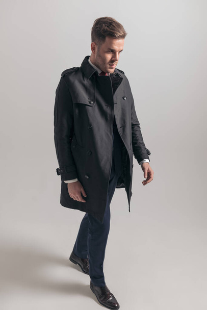 immagine completa del corpo di attraente uomo d'affari in abito e lungo cappotto cercando di lato e camminando con fiducia su sfondo grigio in studio - Foto, immagini