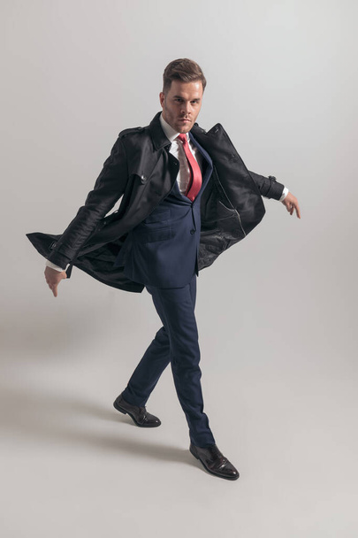 крутой бизнесмен в костюме с длинным пальто, уверенно шагающий перед серым фоном в студии, полное тело - Фото, изображение