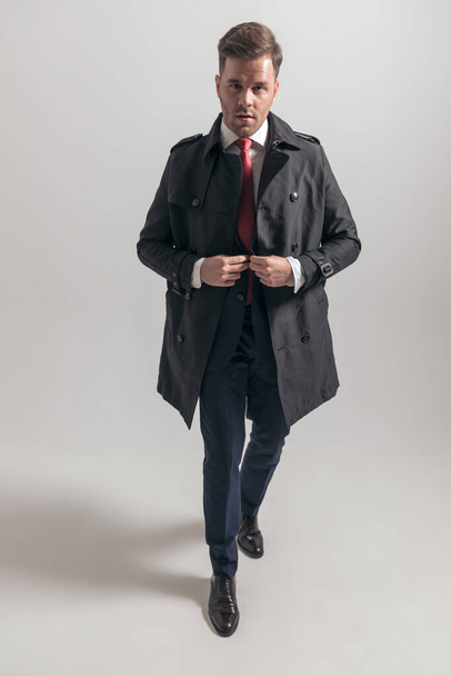 full body picture of elegant businessman with long coat buttoning suit and posing on grey background in studio - Photo, Image