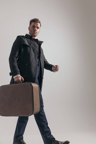 boční pohled na elegantní mladý podnikatel s kufrem chůze na šedém pozadí ve studiu - Fotografie, Obrázek