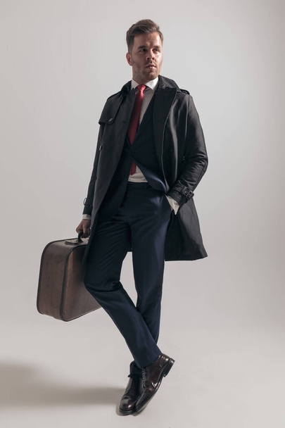 celý obrázek sexy podnikatel v obleku s dlouhým kabátem a kufříkem drží ruku v kapse a dívá se na stranu na šedém pozadí ve studiu - Fotografie, Obrázek