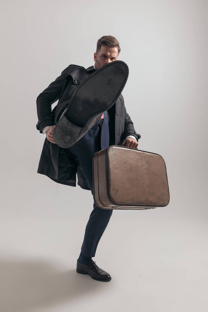 jeune homme d'affaires avec valise tenant la jambe en l'air et se battant sur fond gris en studio - Photo, image