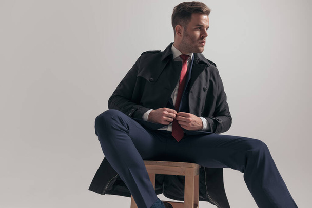 hombre de negocios sentado cerrando traje y mirando a un lado mientras está sentado en la silla de madera y posando sobre fondo gris en el estudio - Foto, Imagen