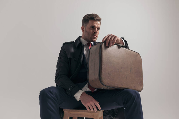 élégant homme d'affaires tenant une valise sur la cuisse, regardant de côté tout en étant assis sur fond gris en studio - Photo, image