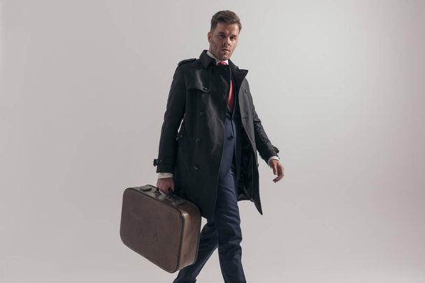 elegancki młody człowiek w garniturze trzymając walizkę i spacerując na szarym tle w studio - Zdjęcie, obraz