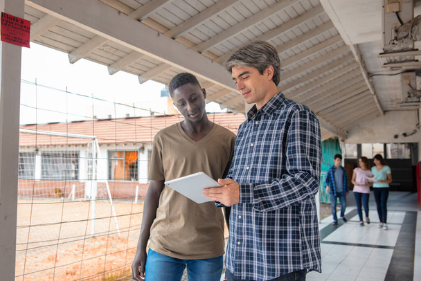 Hight School Afrikalı öğrenci koridorda öğretmeniyle konuşuyor, diğer öğrenciler arka planda konuşuyor. - Fotoğraf, Görsel
