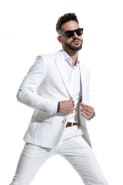 portrait of cool businessman with sunglasses closing and buttoning white suit, posing in a studio fashion light on white background - Fotografie, Obrázek