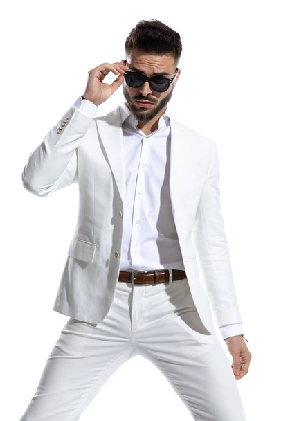 σέξι νεαρός άνδρας με λευκό κοστούμι που διορθώνει γυαλιά ηλίου και ποζάρει σε ένα στούντιο μόδας φως σε λευκό φόντο - Φωτογραφία, εικόνα