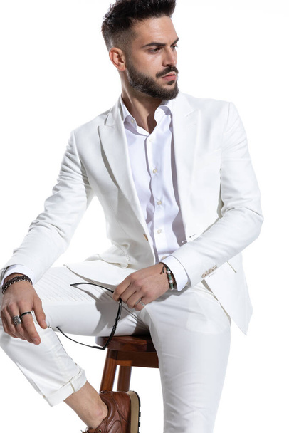 aantrekkelijke zakenman in elegante pak op zoek naar kant en het houden van glazen, terwijl poseren op een witte achtergrond in de studio - Foto, afbeelding
