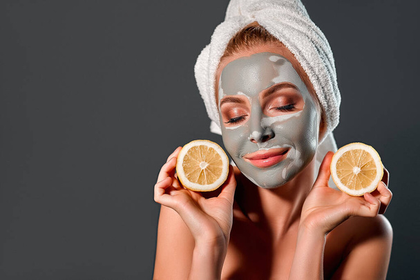 Beauty portrait of a beautiful young woman with gray clay mask on a gray background, holds pieces of lemon near face. - Foto, afbeelding