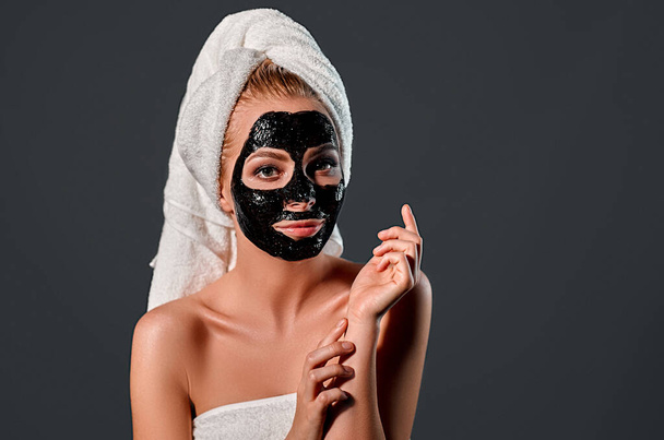 Portrait of a young attractive woman with a towel on her head with a cleansing black mask on her face isolated on a gray background. - Foto, Imagem
