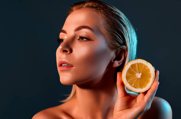 A beautiful woman in profile with perfect skin and bare shoulders on a gray background holding a lemon. - Фото, изображение