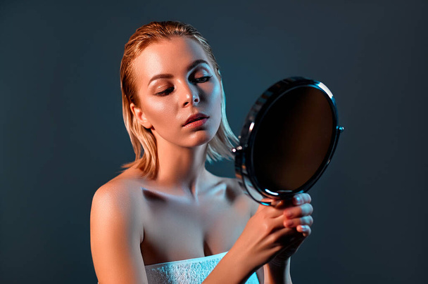 A beautiful girl with blond hair, looking in the mirror at a clean fresh skin. Gray background. Beauty concept. - Fotoğraf, Görsel