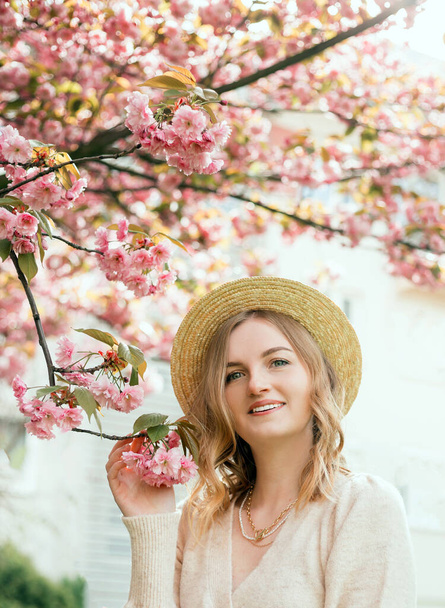 Young stylish woman standing in sakura park and enjoy beauty of pink. Spring woman in cherry flower bloom concept. Beauty blonde woman with long hair outdoors. Cherry blossoms. Close up portrait. - Photo, Image