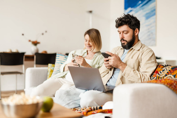 Fehér pár használja mobiltelefonok és laptop, miközben ül a kanapén otthon - Fotó, kép