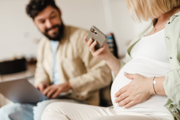 Homem barbudo e mulher grávida usando celular e laptop enquanto sentado no sofá em casa - Foto, Imagem