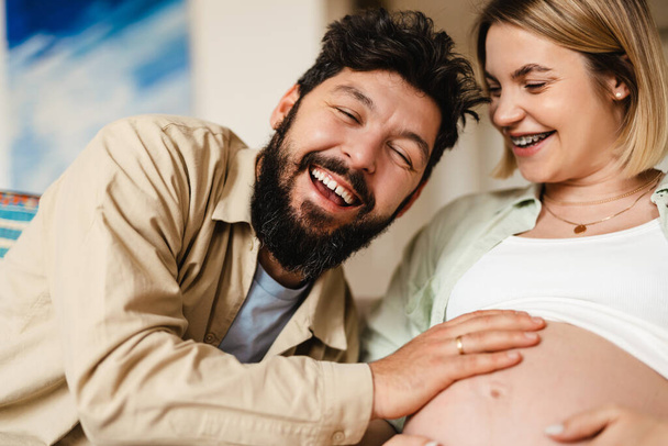 White man touching belly of his pregnant wife while sitting on couch at home - Φωτογραφία, εικόνα